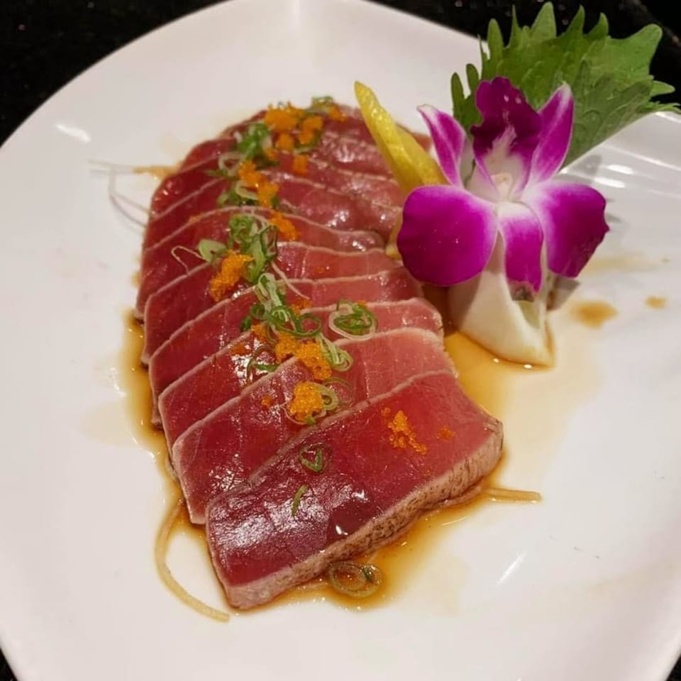 Seared tuna sashimi with green onions, orange tobiko, and a purple orchid garnish on a white plate.