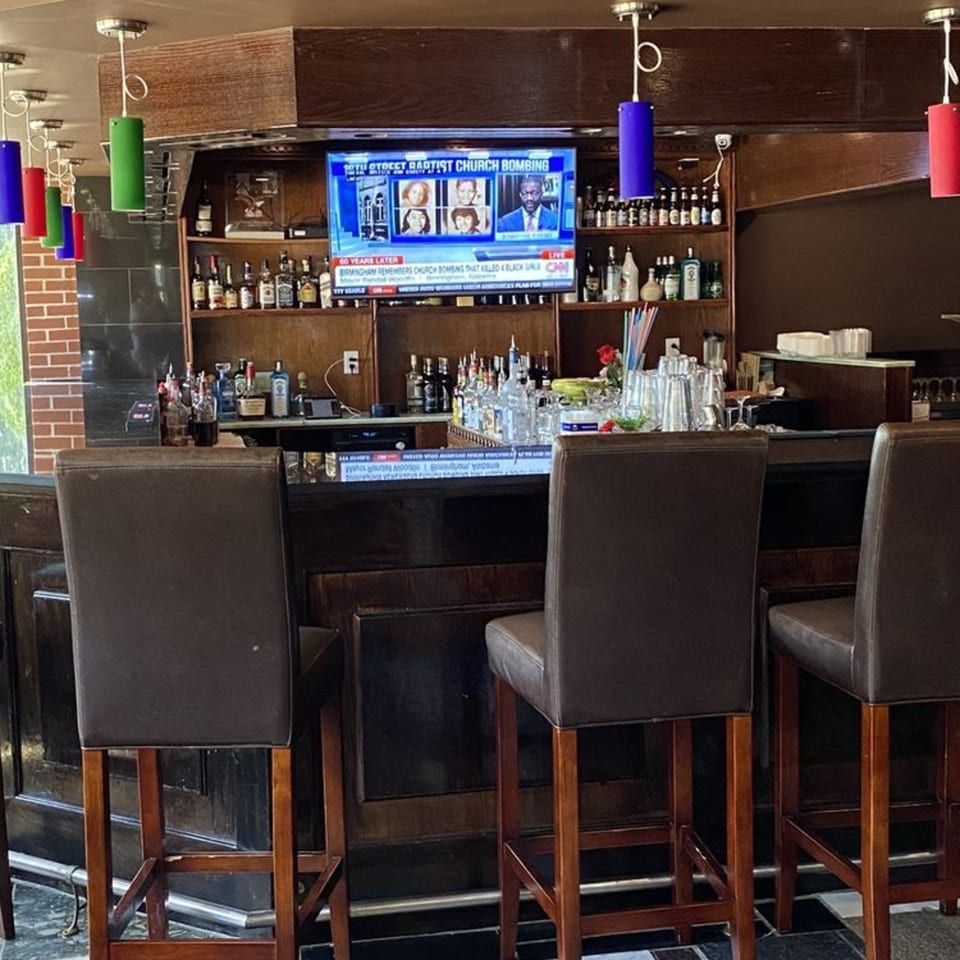 A dark wooden bar with three brown leather stools, liquor bottles, and a TV showing a news report.
