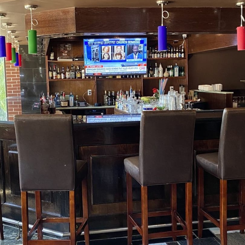 A dark wooden bar with three brown leather stools, liquor bottles, and a TV showing a news report.